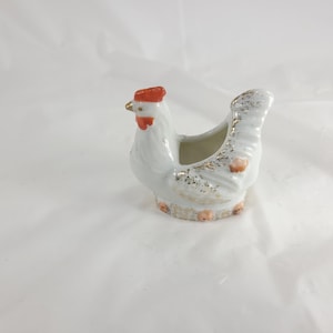May include: A white ceramic hen-shaped container with a red comb and gold accents. The container has a small opening on its back and is decorated with gold flecks and small peach-colored details. The hen is standing on a white surface.