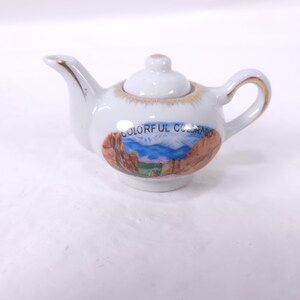 May include: A small white ceramic teapot with gold trim on the spout and handle. The teapot features a colorful landscape scene with the text "COLORFUL COLORADO". The lid is white with a small gold accent.