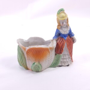May include: A vintage porcelain planter featuring a woman in a blue and red dress, standing next to a tulip-shaped container. The planter has a white, orange, and green color scheme, with a detailed floral design. The woman has blonde hair and a yellow hat.