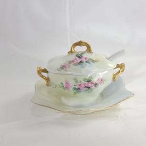 May include: A vintage porcelain sugar bowl with a lid, spoon, and saucer. The bowl and lid are decorated with pink roses and green leaves on a pale yellow background. Gold accents adorn the handles and rim.