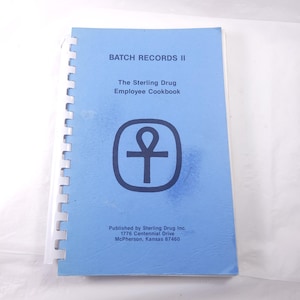 May include: A light blue spiral-bound book titled "BATCH RECORDS II" with the text "The Sterling Drug Employee Cookbook." The cover features a black symbol within a rounded square. The book is published by Sterling Drug Inc.