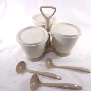 May include: A beige Tupperware serving set with three round containers, each with a lid. The containers are held together by a handle. Three beige ladles are in front of the containers. The set is made of plastic.