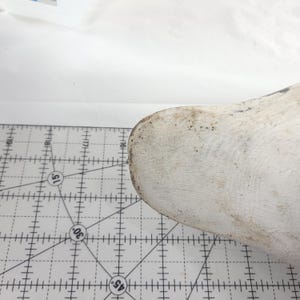 May include: A white wooden shoe last with a worn, textured surface. The shoe last is positioned on a white cutting mat with a grid pattern and degree markings.
