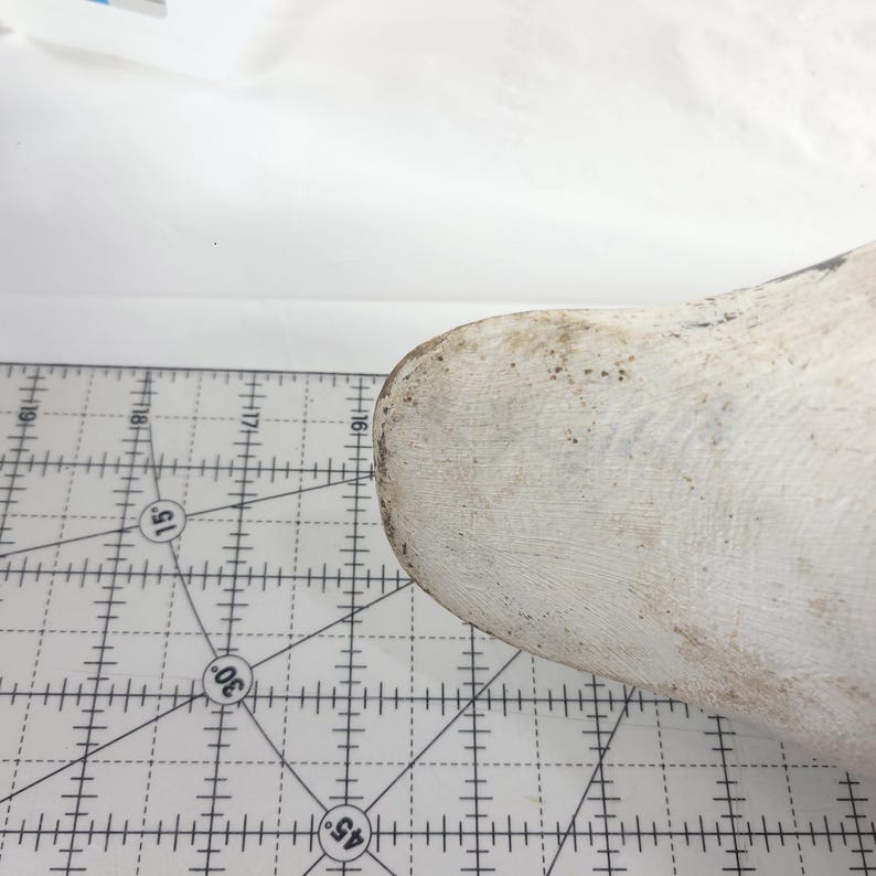May include: A white wooden shoe last with a worn, textured surface. The shoe last is positioned on a white cutting mat with a grid pattern and degree markings.