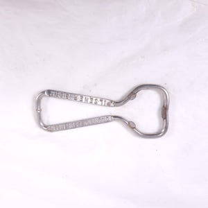 May include: A vintage metal bottle opener with the text "COUNTRY CLUB BEER" and "PILSENER BEER" etched into the metal. The opener has a loop handle and a bottle cap removal end. The metal has a slightly worn appearance.