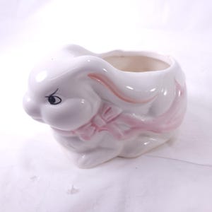 May include: A white ceramic bunny-shaped planter with a pink bow and accents. The bunny has a black eye and pink details on its ears. The planter is designed to hold small plants or decorative items.