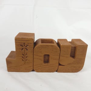 May include: A brown, wood-textured decorative sign spelling the word JOY in block letters. The letter J features a carved flower design on its front face. The letters are connected and stand upright, displaying a consistent grain pattern throughout.