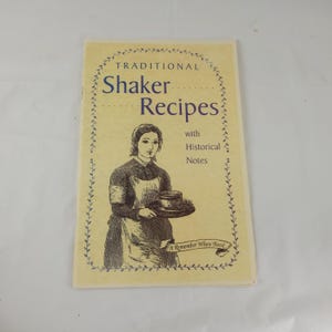 May include: A vintage cookbook titled "Traditional Shaker Recipes" with a drawing of a woman holding a tray. The cover is cream-colored with blue text and a decorative border. The book includes historical notes.