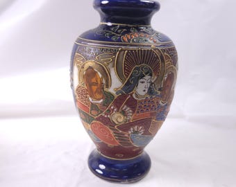May include: A small, blue and white ceramic vase with a Japanese-inspired design. The vase features a scene of three figures in traditional clothing, painted in gold, red, and black.
