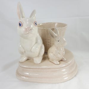 May include: A ceramic Easter-themed decorative piece featuring two white rabbits with blue eyes and a woven basket. The rabbits are positioned on a light beige base, creating a charming and whimsical display.