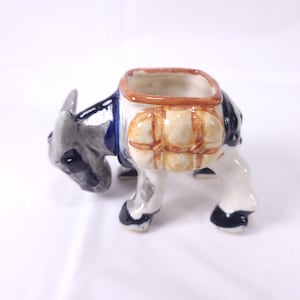 May include: A ceramic donkey figurine with a square opening on its back, painted in shades of gray, white, and black. The donkey has a brown and tan pack saddle. The figurine is a decorative item.