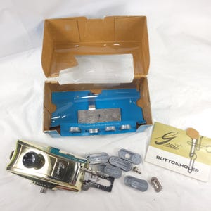May include: A vintage Greist Buttonholer set with a gold-toned metal buttonholer, various gray attachments, and a cardboard box with a blue interior. The set includes an instruction sheet and is designed for sewing.