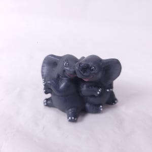 May include: A dark gray figurine of two elephants embracing. The elephants have large ears, small white dots, and pink mouths. The figurine is on a white surface.