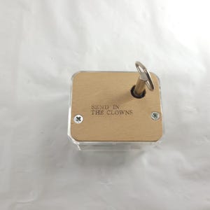 May include: A small, rectangular music box with a clear plastic base and a light wood top. The top has the phrase "SEND IN THE CLOWNS" engraved on it. A silver-colored key is inserted into the top.