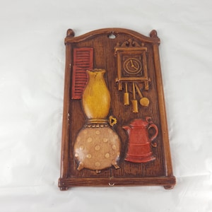 May include: A vintage-style wall plaque with a brown wooden frame. The plaque features relief carvings of a yellow oil lamp, a red teapot, a clock, and a shutter. The lamp has a tan base with brown polka dots. The clock has a circular face.