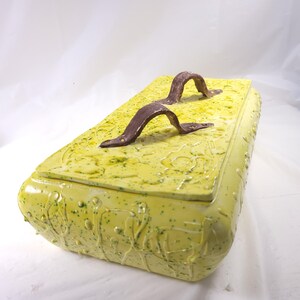 May include: A rectangular ceramic box with a yellow-green glaze and brown handles. The box has a textured surface with embossed patterns and green speckles. The handles are arched and attached with brown hardware.