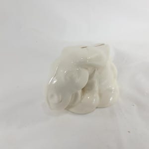 May include: A cream-colored ceramic piggy bank shaped like a cluster of clouds. The bank has two slots on top for inserting coins. The glossy finish gives it a smooth, reflective appearance. The design is simple and elegant.