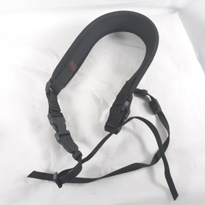 May include: Black camera neck strap with a padded shoulder rest. The strap has adjustable nylon webbing and plastic buckles for secure attachment. A small red tag is visible on the strap.