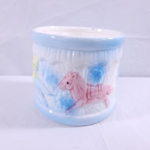 Op de afbeelding: Een witte keramische pot met een blauwe rand en een carrouselscène. De pot heeft een roze paard, een gele vogel en blauwe bloemen.