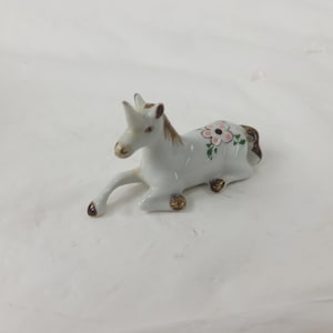 May include: A white ceramic figurine of a reclining horse with brown mane and tail. The horse has a pink floral design on its side and gold accents on its hooves. The figurine is a decorative item.