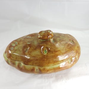 May include: A ceramic lidded serving dish with a brown and green glaze. The dish is oval-shaped with a textured surface and a small knob on the lid. The glaze has a glossy finish, and the dish appears to be handmade.