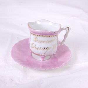 May include: A vintage pink teacup and saucer set. The teacup has a white interior and a pink exterior with gold trim. The saucer is also pink. The teacup is inscribed with the words "Souvenir Chicago Ill" in gold lettering.