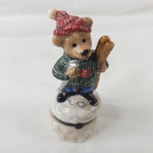 May include: A ceramic figurine of a light brown bear wearing a red knit hat with a pom-pom, a dark green cable-knit sweater, and blue pants. The bear holds wooden skis and stands on a white, textured base featuring a pair of small silver glasses.