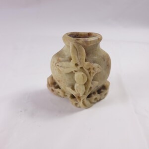 May include: A small, carved stone vase with a light green and brown color. The vase is decorated with a floral design.