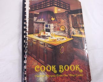 May include: Vintage cookbook with a spiral binding. The cover features a kitchen scene with a brick wall, wooden cabinets, and the title "COOK BOOK" in gold lettering. The subtitle reads "Favorite Recipes From Our Best Cooks."
