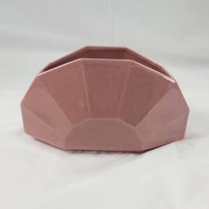 May include: A dusty rose-colored, geometric ceramic container. The container has a faceted design with a textured surface. The open top reveals the interior, and the piece sits on a white surface.
