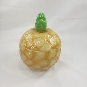May include: A ceramic pineapple-shaped jar with a removable lid. The body of the jar features a textured, golden-brown, diamond-patterned surface mimicking pineapple skin, topped with a glossy, bright green, pointed leaf finial handle.
