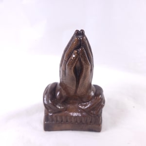 May include: A brown sculpture of hands clasped in prayer. The hands are detailed with visible knuckles and skin texture. The sculpture sits on a rectangular base and is set against a white background.