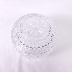 May include: A clear, round glass jar with a diamond-cut pattern. The jar has a lid with a floral design in the center. The jar is sitting on a white surface.