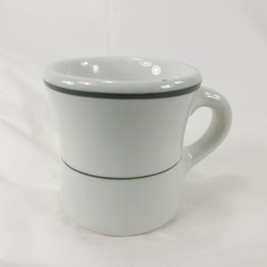 May include: A white ceramic mug featuring a thick, rounded rim and a sturdy handle. The mug is decorated with two thin, dark green horizontal stripes, one encircling the upper portion near the rim and the other positioned around the lower body.