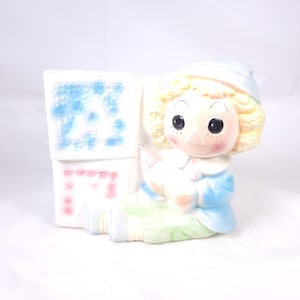 May include: A vintage ceramic baby planter featuring a seated doll figure with blonde curly hair and a blue cap. The doll is holding a heart and is next to white blocks with the letter "A" in blue and pink. The planter has a pastel color scheme.