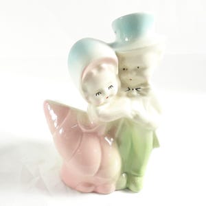 May include: A vintage ceramic figurine of a couple embracing. The woman is wearing a pink and blue dress and the man is wearing a green suit and a blue top hat. The couple is standing on a pink and white base.