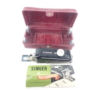May include: A vintage Singer buttonholer sewing machine attachment with a black body and white dial, accompanied by a maroon case and a green and white instruction booklet. The booklet reads "SINGER BUTTONHOLER".