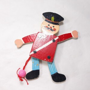 May include: A wooden pull toy of a soldier with a red and white uniform, black boots, and a black hat. The toy has a pink ball attached to a string.