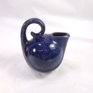May include: A small, navy blue ceramic oil lamp with a curved handle and a spout. The lamp has a speckled texture and a glossy finish. The design is reminiscent of ancient pottery.