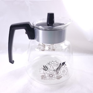 May include: A clear glass coffee carafe with a black handle and lid. The carafe has a silver band around the middle and is decorated with a floral pattern in shades of gray and white. The carafe is designed to hold hot liquids.