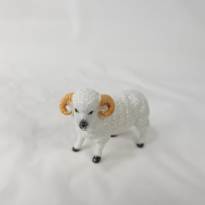 May include: A small ceramic figurine of a ram with a white textured body, curved golden-brown horns, and black painted details on the nose and hooves. The ram is standing in a forward-facing position with its head slightly turned to the left side.