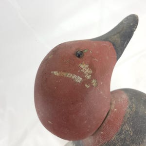 May include: Close-up of a vintage wooden duck decoy. The duck's head is painted a faded red with a black beak and a small, dark eye. The paint shows signs of wear, adding to its antique charm. The neck is also painted red.