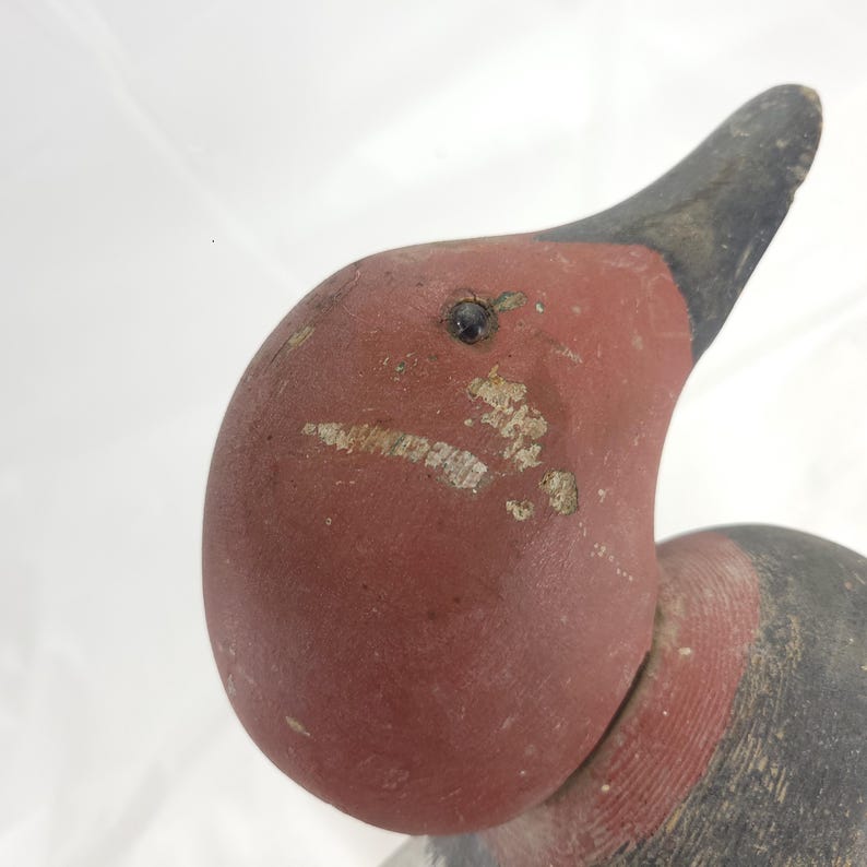 May include: Close-up of a vintage wooden duck decoy. The duck's head is painted a faded red with a black beak and a small, dark eye. The paint shows signs of wear, adding to its antique charm. The neck is also painted red.