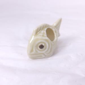May include: A ceramic fish-shaped object in a light cream color. The fish has a detailed design with a brown eye and an open mouth. The object is likely a decorative item or a small container.