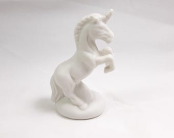 May include: White ceramic unicorn figurine standing on a round base. The unicorn is rearing up on its hind legs with its horn pointing upwards.