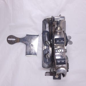 May include: A vintage silver-toned sewing machine attachment with a lever and a small, separate metal piece. The attachment has a series of screws and black rubber pads. The word "Ramona" is visible on the side. The item is likely used for sewing or fabric manipulation.