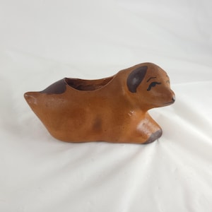 May include: A brown ceramic dog-shaped planter. The planter has a light brown body with darker brown accents on the ears, tail, and paws. The dog is in a resting position, with a small opening for planting.