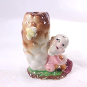 May include: A vintage ceramic figurine featuring a small child leaning against a tree trunk. The tree trunk is brown and white, with a yellow and green bee attached. The child is wearing a pink outfit and has a red basket.