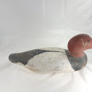 May include: A vintage wooden duck decoy, painted in shades of white, black, and red. The decoy features a detailed design with a rounded head and a long, streamlined body. The duck is likely used for hunting or decorative purposes.