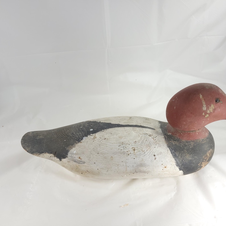 May include: A vintage wooden duck decoy, painted in shades of white, black, and red. The decoy features a detailed design with a rounded head and a long, streamlined body. The duck is likely used for hunting or decorative purposes.